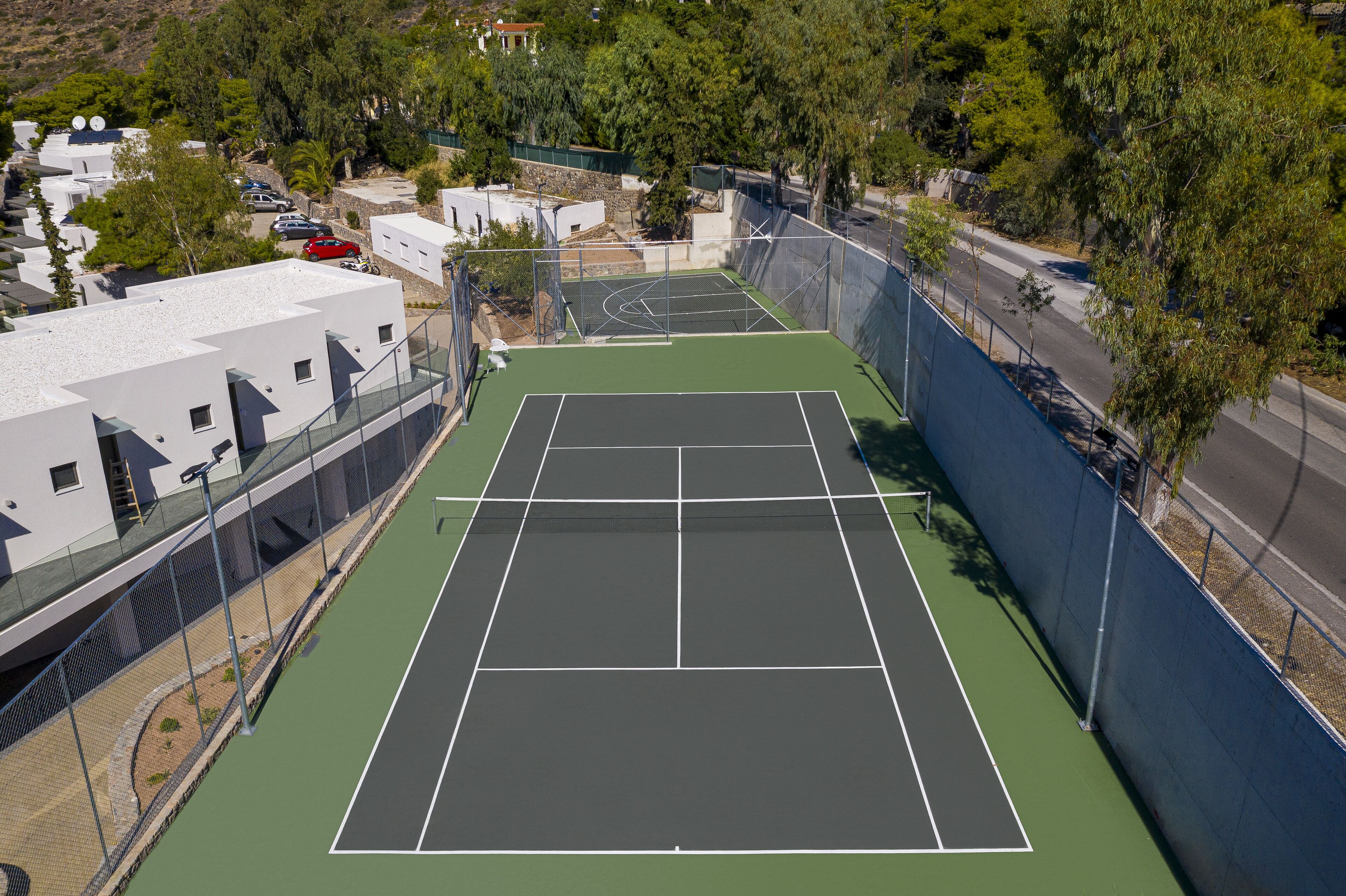 An outdoor tennis court surrounded by a blue fence, with a road and trees in the background.