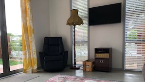 Ein Zimmer mit einem schwarzen Liegesessel, einem Holzschrank, einer Lampe und einem großen Fenster mit Jalousien.