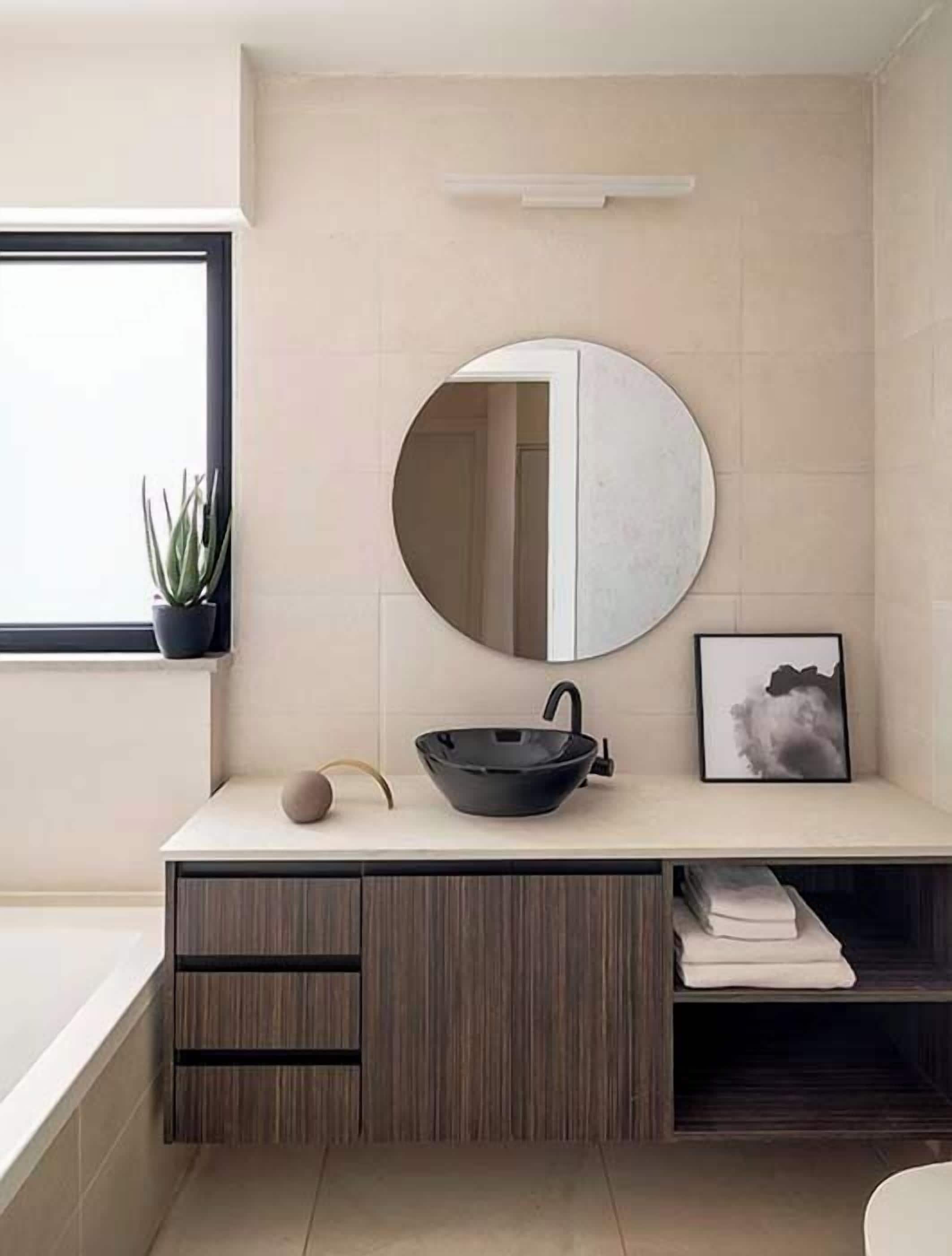 A modern bathroom with a round mirror, black vessel sink, wooden vanity, and a potted plant.