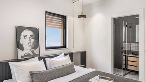 A modern bedroom with a bed, a window with blinds, a bedside table with a book, and a bathroom visible through an open door. - Mr. Louis - By TLV2GO (Tel Aviv)