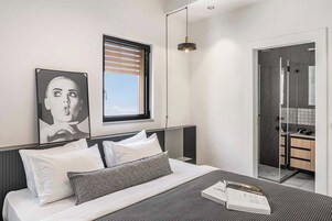 Ein modernes Schlafzimmer mit Bett, einem Fenster mit Jalousien, einem Nachttisch mit Buch und einem durchgehenden Türspalt sichtbaren Badezimmer.
