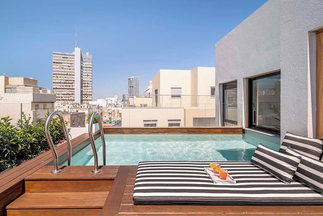 Uma área de piscina no terraço com uma espreguiçadeira e uma mesa auxiliar, com vista para um horizonte urbano repleto de edifícios modernos.