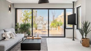 A modern living room with a sofa, coffee table, and TV, large sliding glass doors to a balcony with plants and city views. - Mr. Louis - By TLV2GO (Tel Aviv)