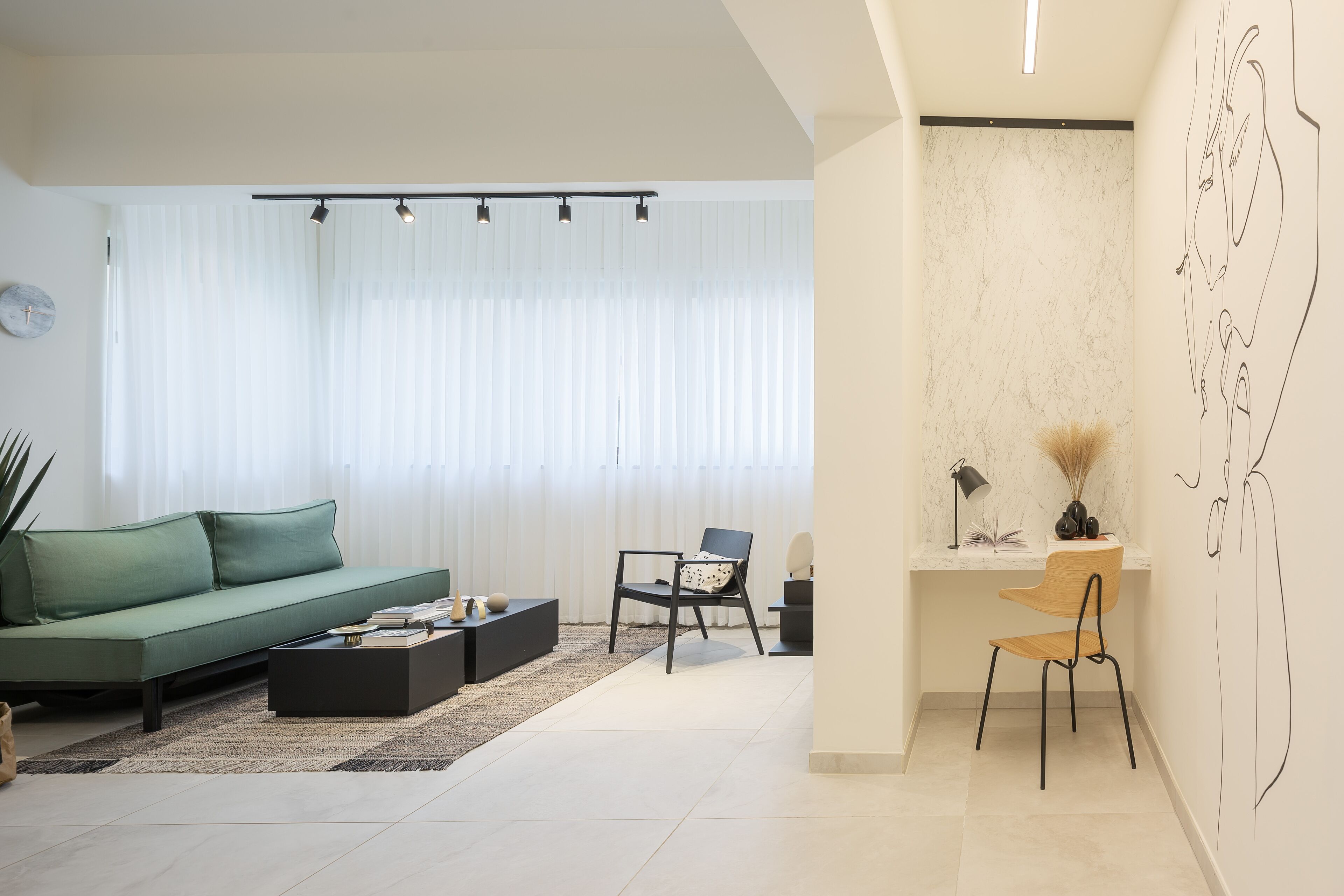 A modern living room with a green sofa, a black coffee table, and a wooden chair. There is a desk with a lamp and a vase of dried plants. The room has large windows with sheer curtains and a marble wall with a line drawing.