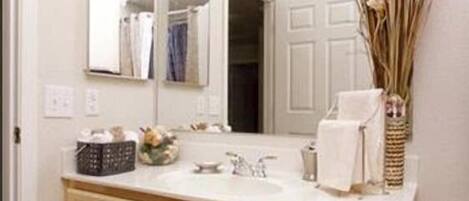 A bathroom with a double vanity, a mirror with lights, a vase with dried plants, and a towel rack.