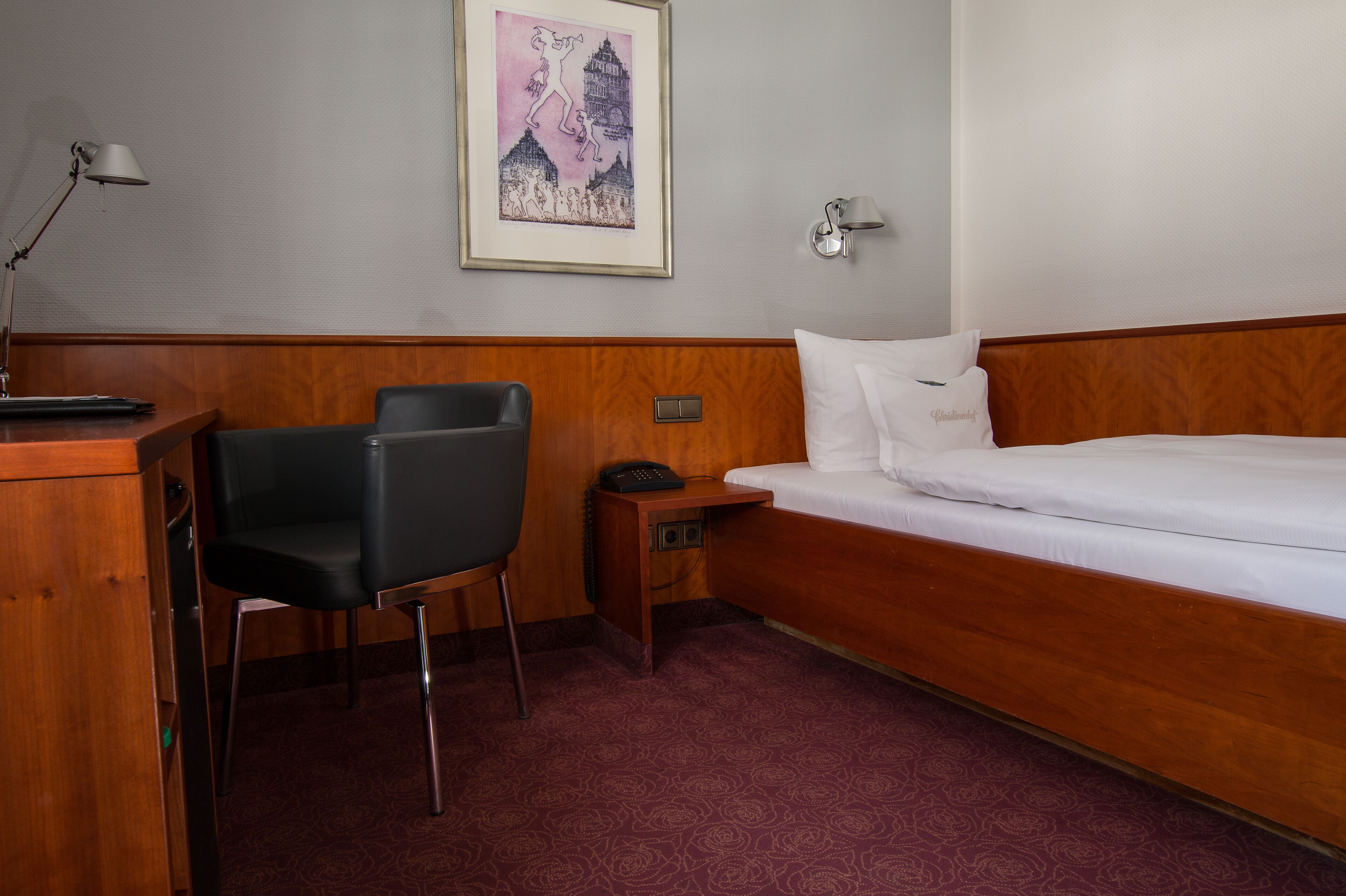 A hotel room with a wooden headboard, a single bed, a desk with a chair, a nightstand, and a wall-mounted lamp.