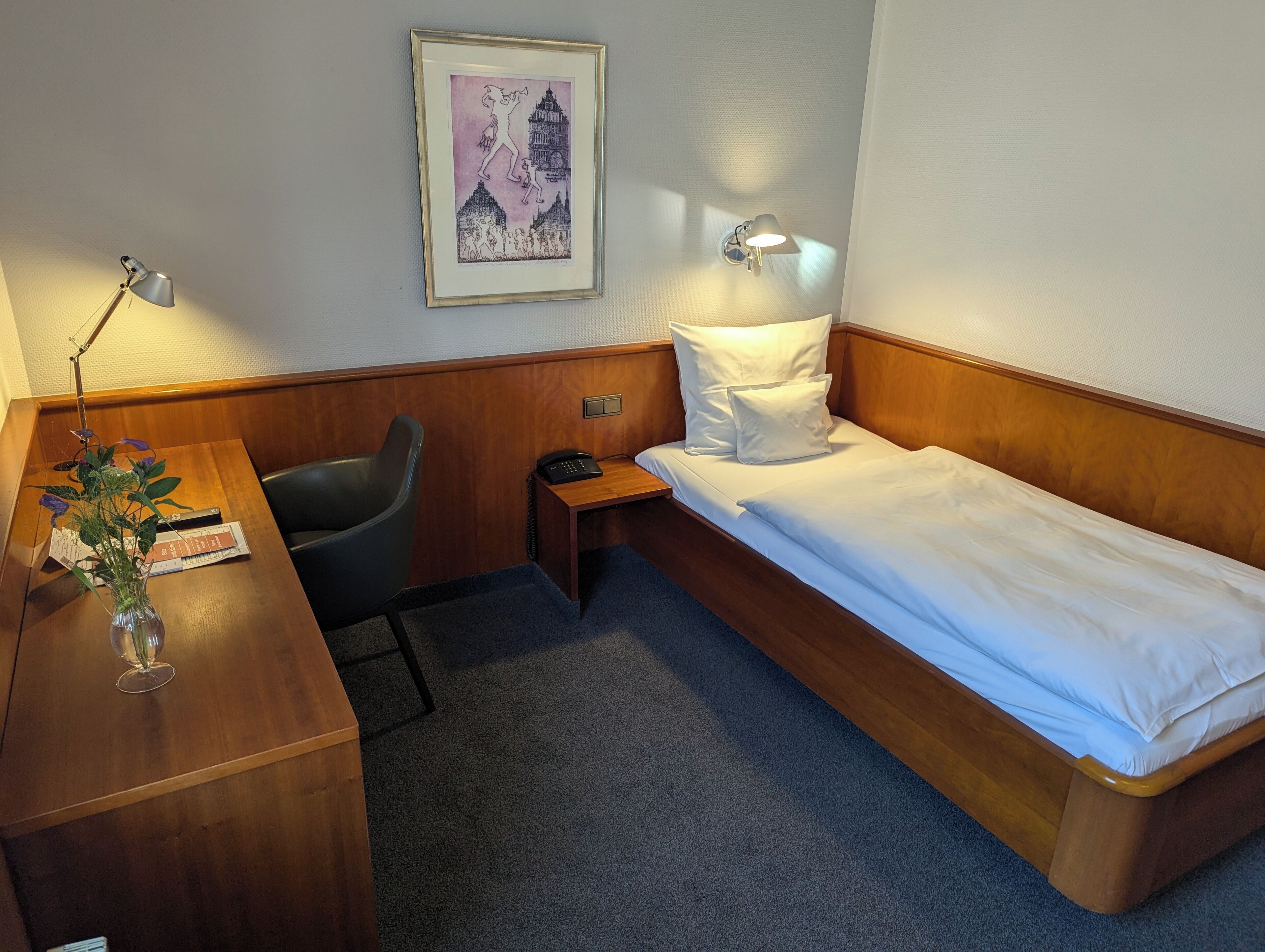 Habitació d'hotel amb escriptori de fusta, una cadira, una petita taula de nit amb una làmpada, un llit amb roba blanca i un quadre al mur.
