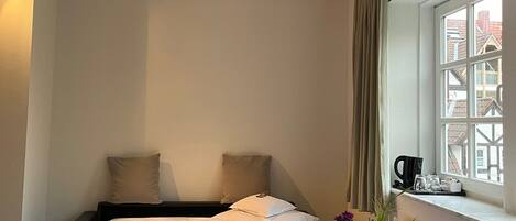 Habitación de hotel con cama, mesita de noche con un florero, ventana con cortinas y vistas a edificios.