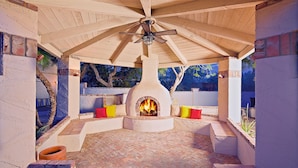A covered outdoor patio with a brick floor, a central fire pit, and seating areas with cushions. - Arizona Desert Haven (Scottsdale)