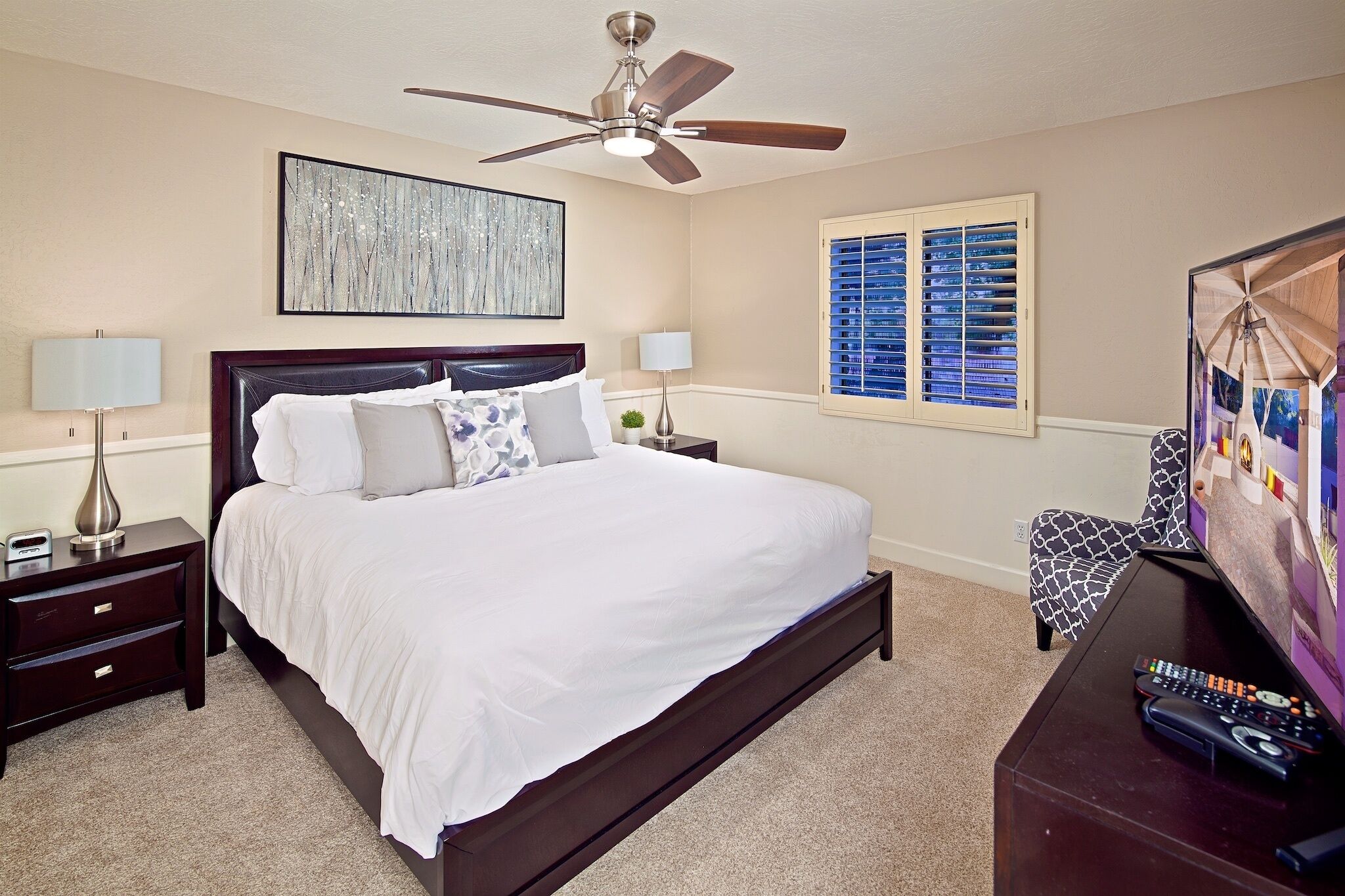 A bedroom with a bed, nightstands, a ceiling fan, a window with blinds, and a TV.