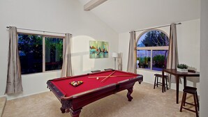 A pool table in a room with large windows, a painting on the wall, and a table with chairs. - Tatum Place (Phoenix)