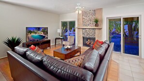 A modern living room with a leather sofa, a coffee table, a TV, a fireplace, and a view of a pool through large sliding glass doors. - Tatum Place (Phoenix)