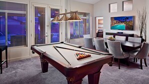 A pool table in a room with a dining area, a TV mounted on the wall, and large windows with blinds. - Kierland Retreat (Scottsdale)