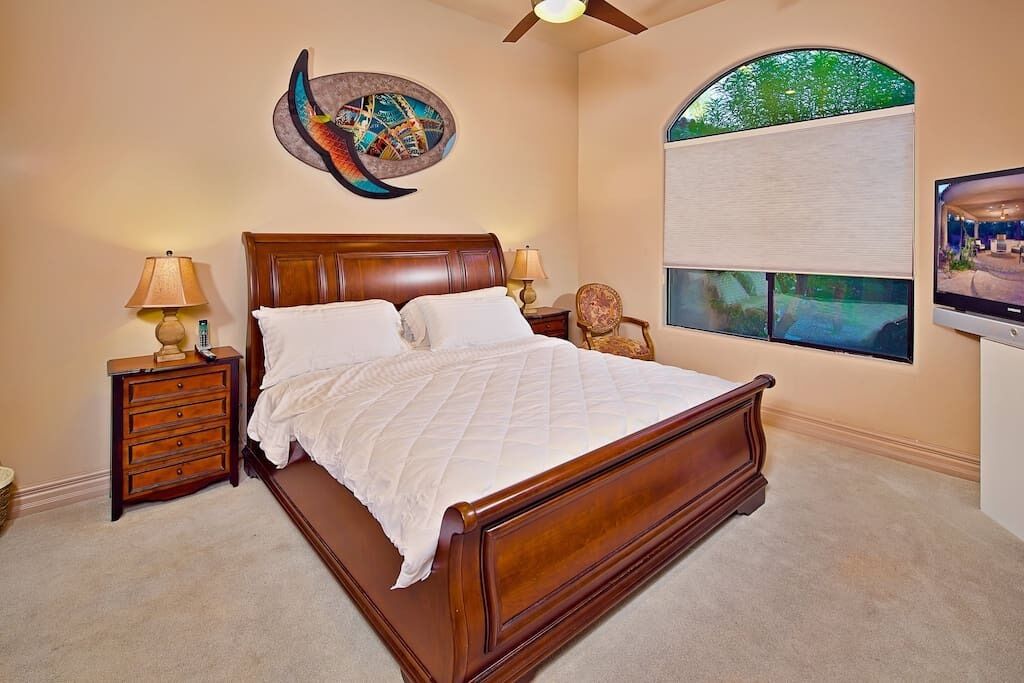 Un dormitorio con cama de madera, dos mesitas de noche con lámparas, un reloj de pared, un televisor y un amplio ventanal con vistas a zonas verdes.