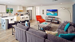 A modern living room with a grey sectional sofa, a red armchair, and a coffee table. A flat-screen TV is mounted on the wall, and there is a kitchen area visible in the background. - Old Town Retreat (Scottsdale)