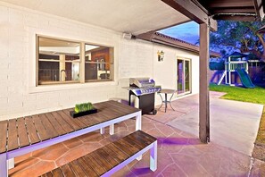 Um pátio externo com mesa e banco de madeira, churrasqueira e área de recreação ao fundo.