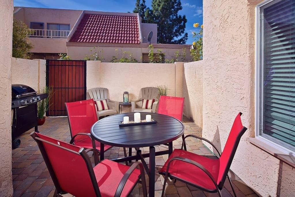 A patio with a round table and red chairs, a grill, and a view of a house with a red-tiled roof.
