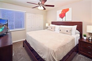 A bedroom with a bed, a ceiling fan, a TV, a window with blinds, a door, and a painting. - Silverado Townhouse (Scottsdale)