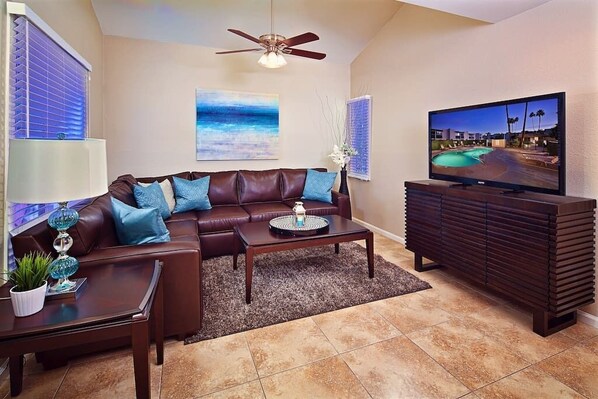 A living room with a brown leather sofa, a coffee table, a TV, and a ceiling fan. - Silverado Townhouse (Scottsdale)