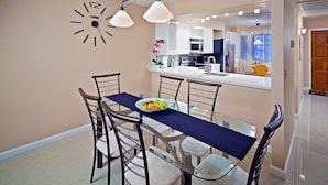 A dining area with a glass table, metal chairs, a bowl of fruit, and a wall clock. - Tempe Escape · Prime Location, Sparkling Pool, Bbq, Tennis Court (Tempe)