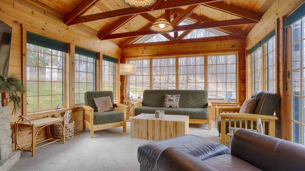 A cozy cabin interior with wooden walls and a high ceiling. There are two sofas, a coffee table, and a wooden bench. Large windows provide views of the surrounding forest.