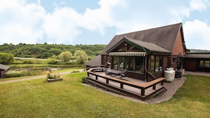 A log cabin with a deck, a pond, and a forested hill in the background.