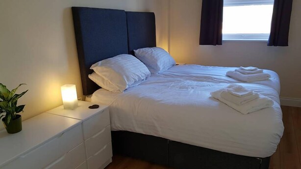 Una cama bien hecha con sábanas blancas y toallas dobladas, una lámpara de noche, una planta en maceta y un cabecero oscuro en una habitación de hotel con suelo de madera.