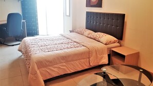 Un dormitorio con cama, escritorio, silla, mesa de vidrio y un cuadro en la pared.
