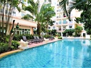 A swimming pool with lounge chairs and palm trees, surrounded by a multi-story building with balconies. - Park Lane studio apartment with swimming pool view Pattaya (เมืองพัทยา)