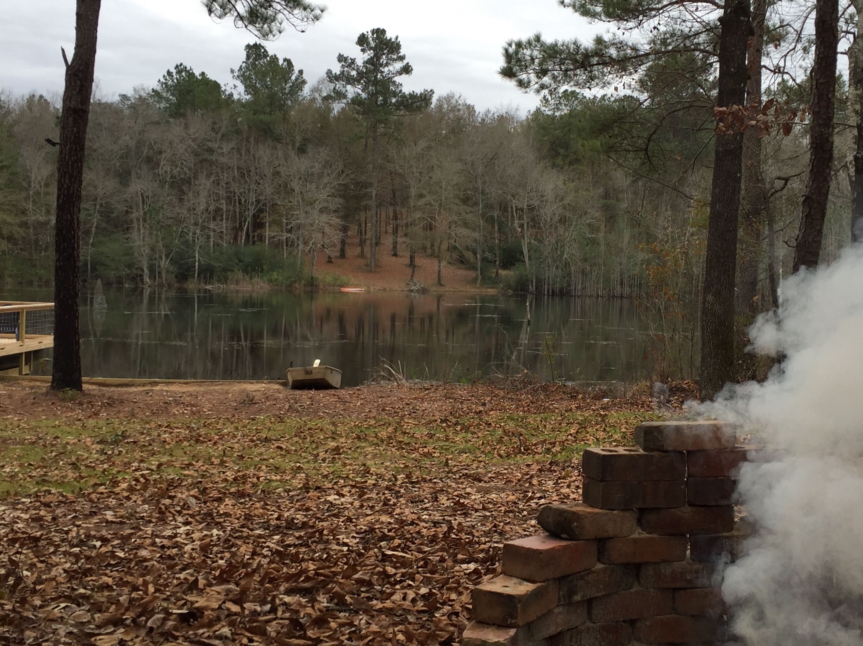 A serene outdoor setting with a wooden dock, a small boat, and a brick structure emitting smoke, surrounded by a forested area and a calm lake.