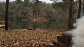 A serene outdoor setting with a wooden dock, a small boat, and a brick structure emitting smoke, surrounded by a forested area and a calm lake.