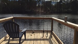 A wooden deck with a chair and fishing rods, overlooking a calm lake with trees in the background.