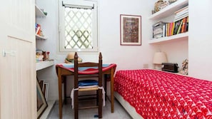 Ein Schlafzimmer mit Bett, Schreibtisch, Stuhl, Fenster, Bücherregal und einem gerahmten Bild an der Wand.
