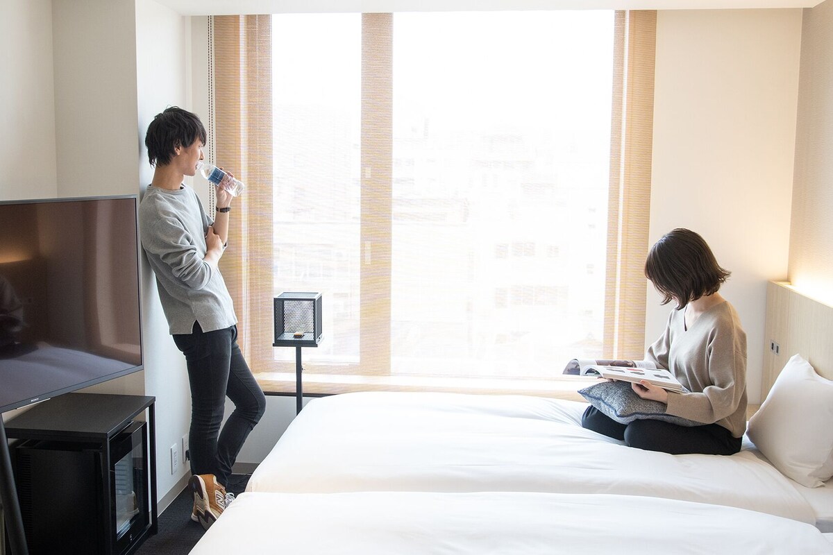 A hotel room with a bed, a TV, a window with blinds, and a small table with a lamp.