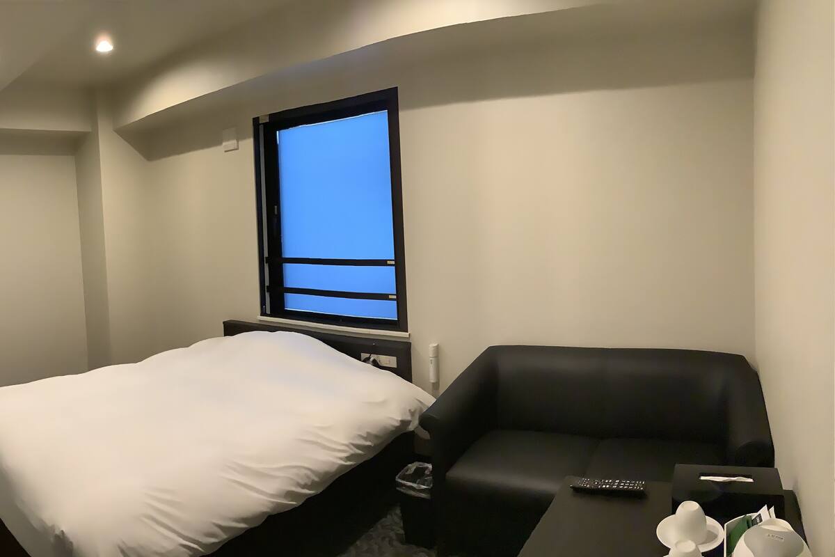 A hotel room with a bed, a black sofa, a window with a blue view, and a remote control on the sofa.