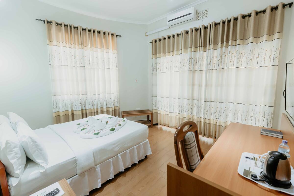 Une chambre d’hôtel avec un lit, un bureau en bois, une chaise, une table, une bouilloire et des rideaux.