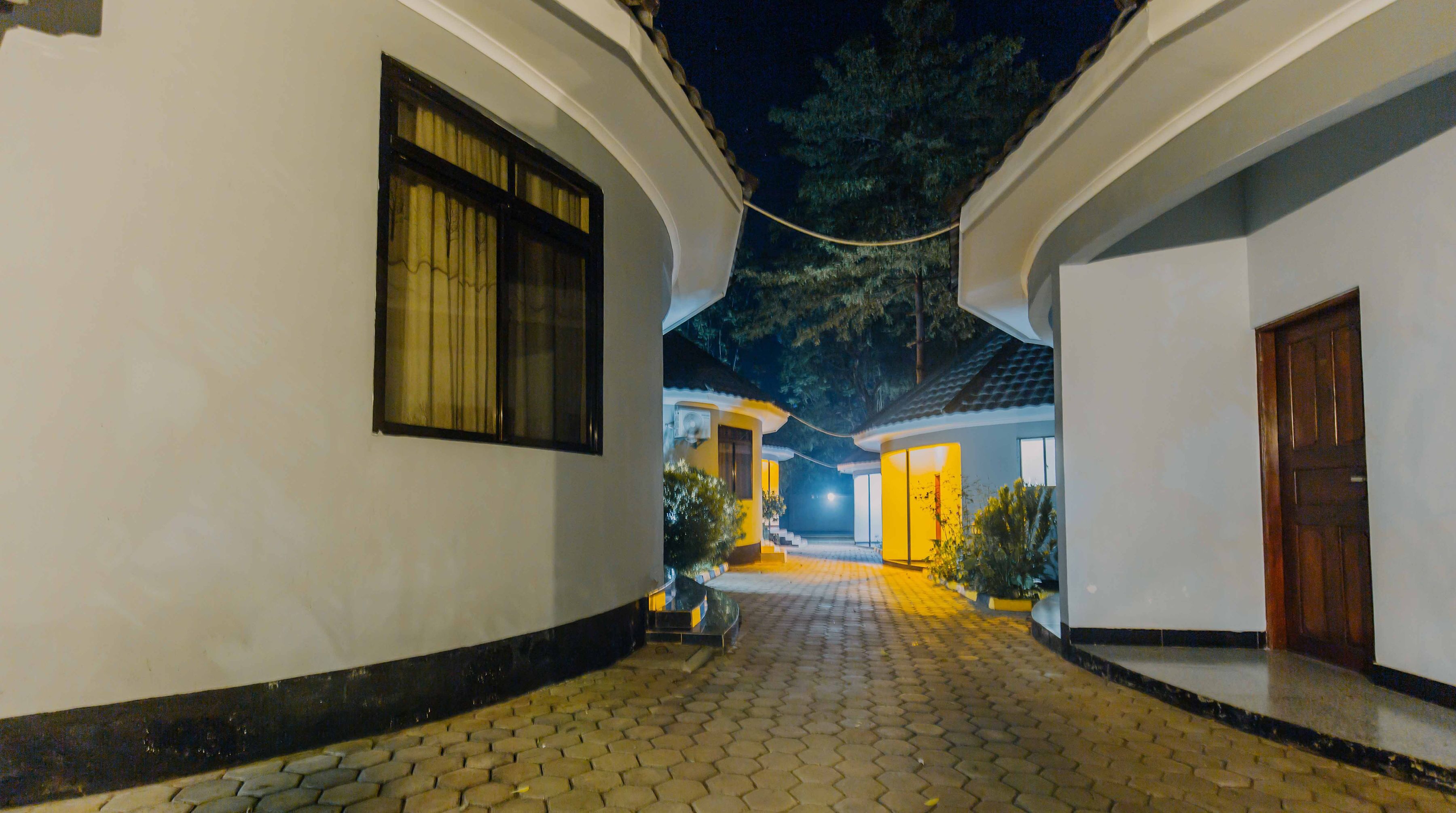 A nighttime view of a courtyard with a paved walkway, a building with a large window, and a door.