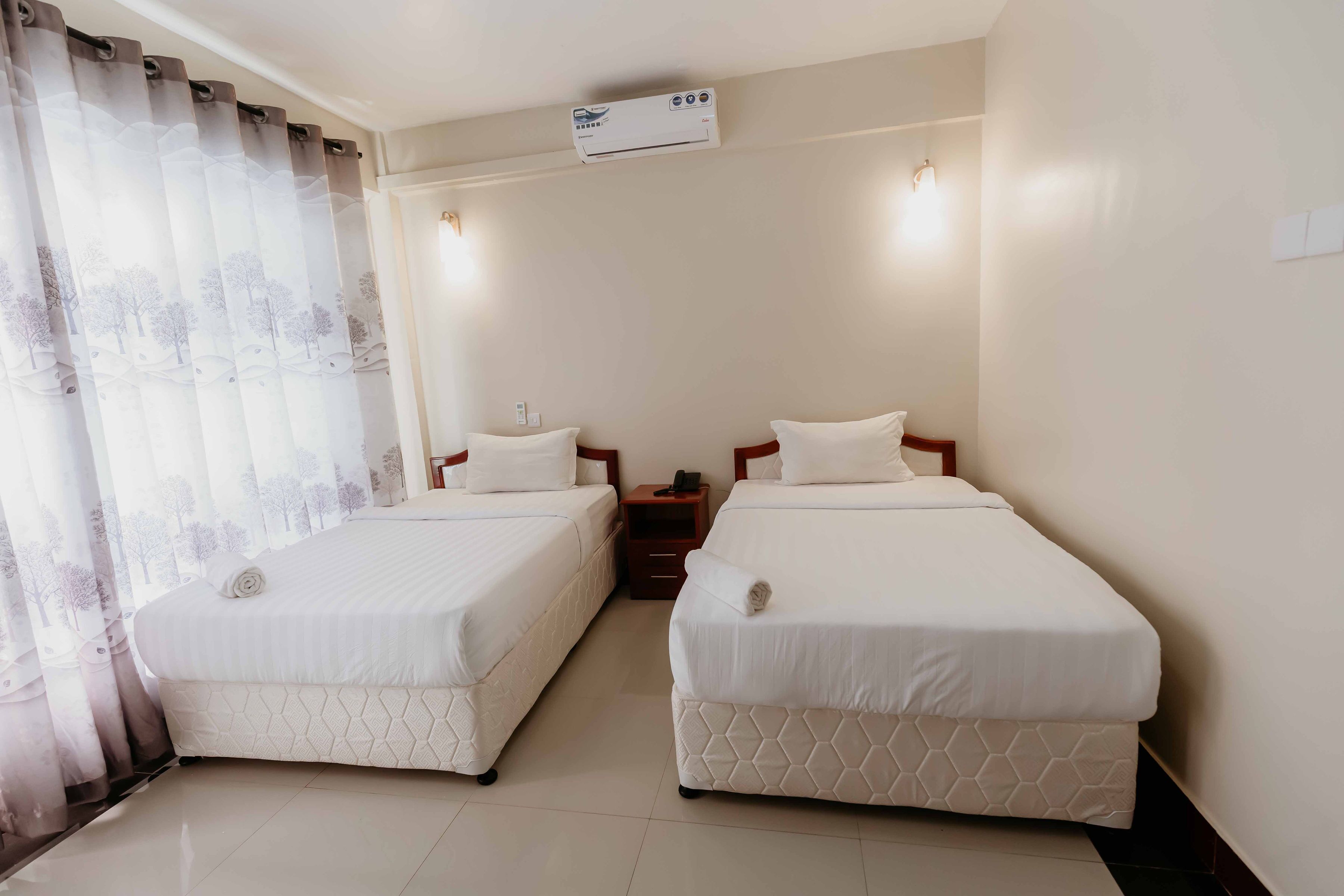 A hotel room with two single beds, a nightstand, an air conditioning unit, and a window with sheer curtains.