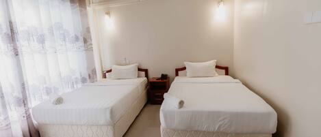 Habitación de hotel con dos camas individuales, una mesita de noche, un equipo de aire acondicionado y una ventana con cortinas translúcidas.
