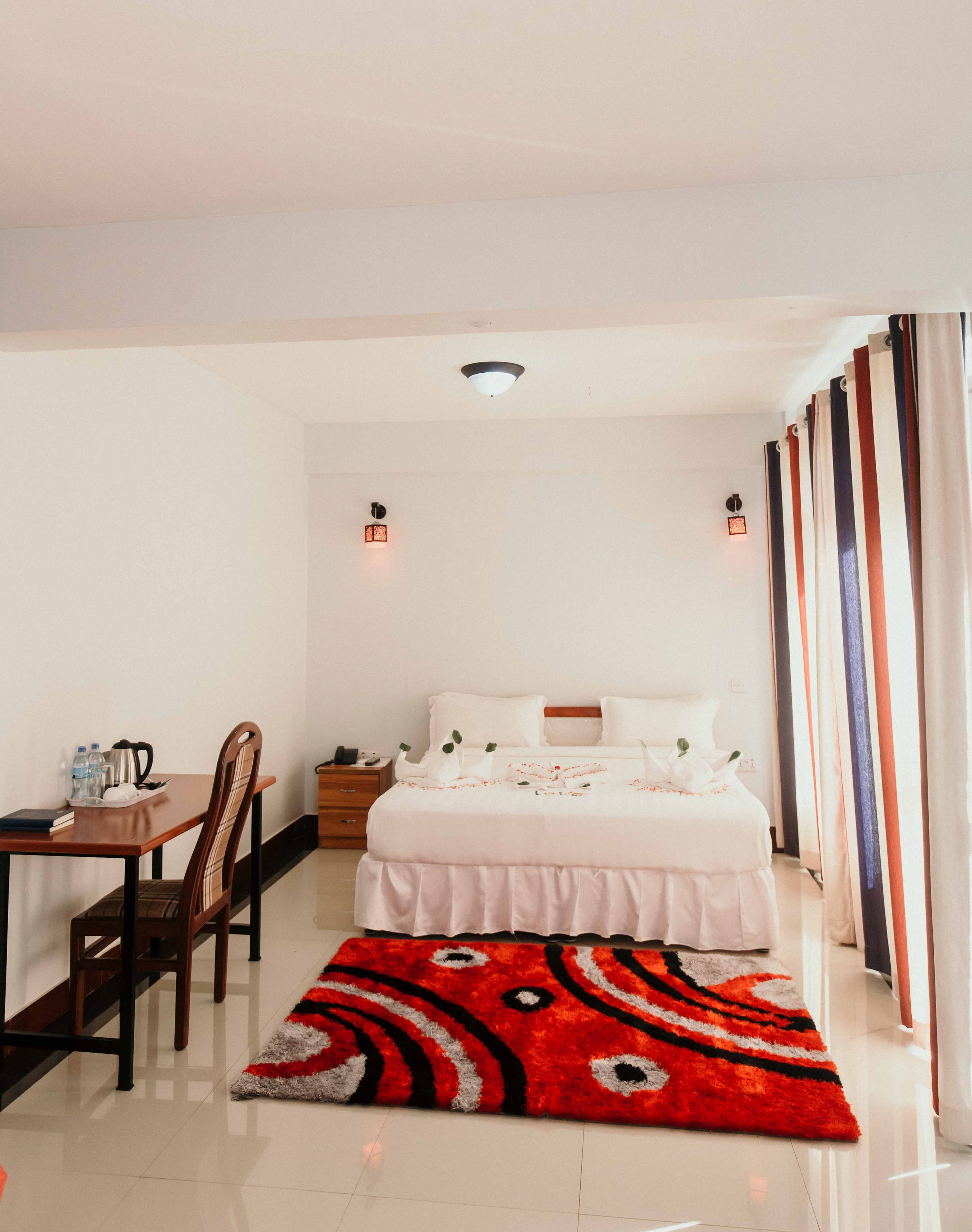 Habitación de hotel con cama, escritorio, silla, mesita de noche y alfombra roja estampada.