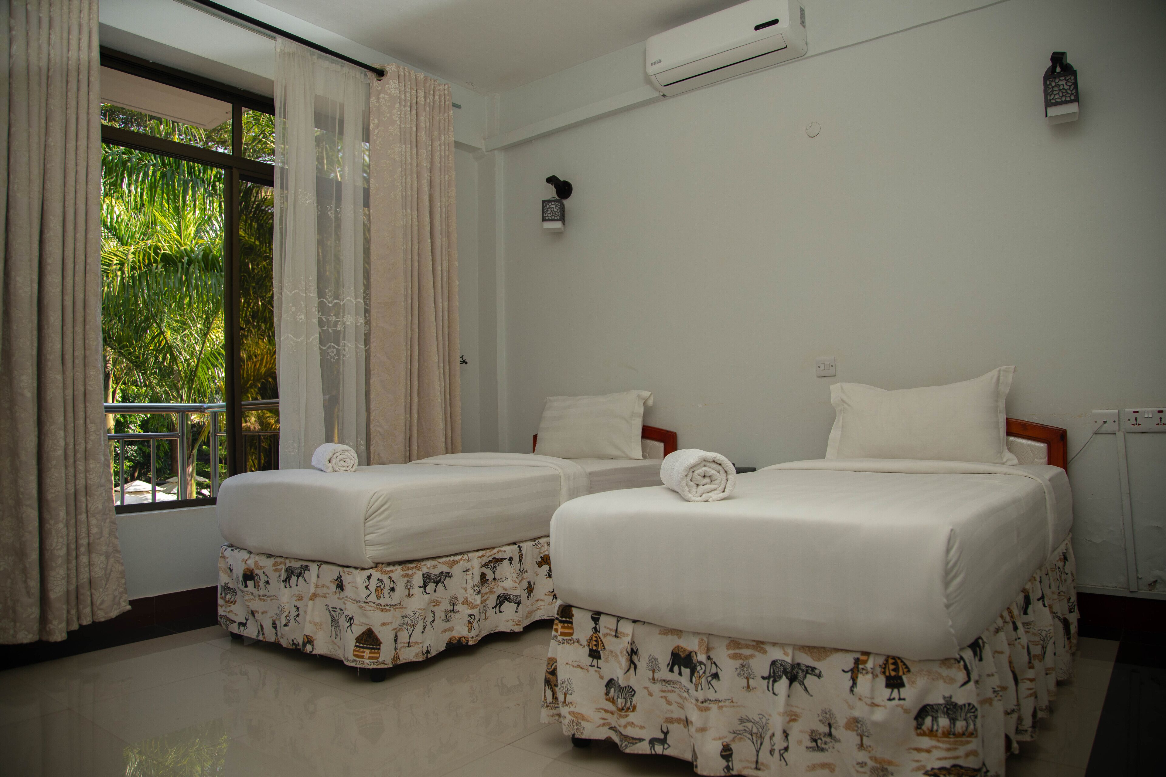 A hotel room with two beds, a window with curtains, an air conditioning unit, and a small wall-mounted light fixture.