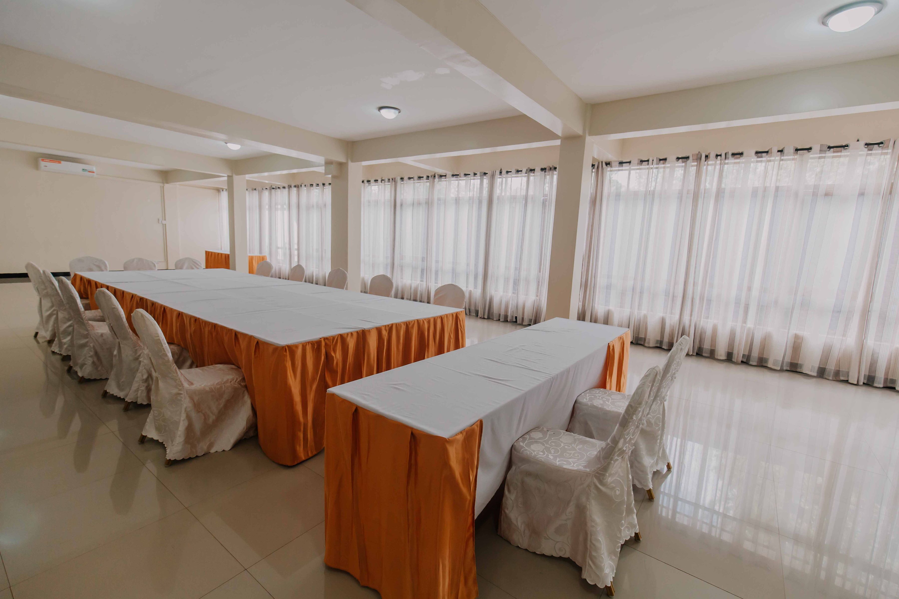 Uma sala de conferências com uma mesa comprida e cadeiras, janelas amplas com cortinas e piso ladrilhado.