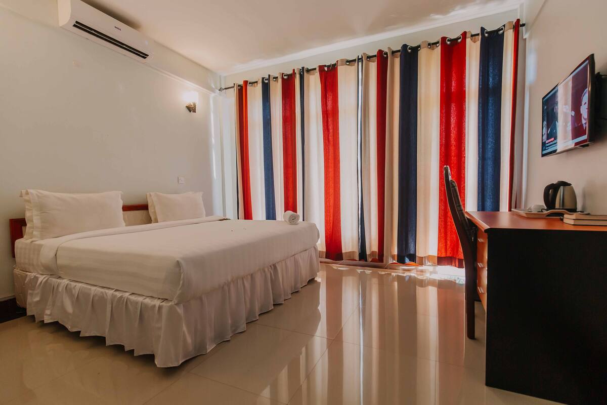 Une chambre d’hôtel équipée d’un lit, d’un bureau, d’une chaise, d’une télévision et d’une fenêtre avec des rideaux à rayures.