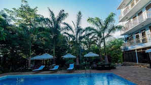 A swimming pool with lounge chairs and umbrellas, surrounded by palm trees and a building with balconies. - Panama Garden Resort (Moshi)