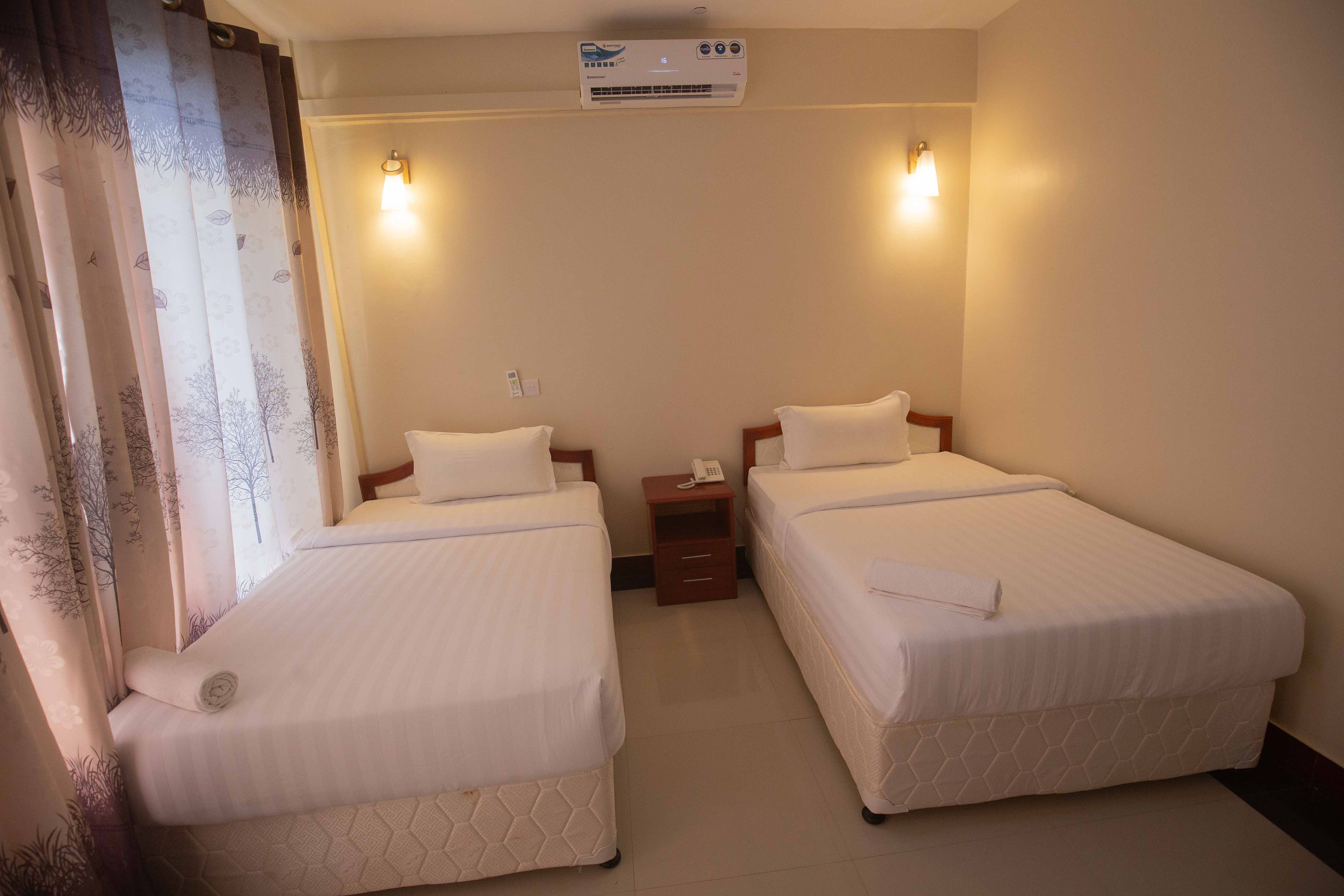 A hotel room with two beds, a bedside table with a phone, and an air conditioning unit on the wall.