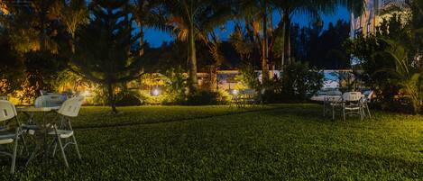 A well-lit outdoor seating area with tables and chairs, surrounded by lush greenery and palm trees.