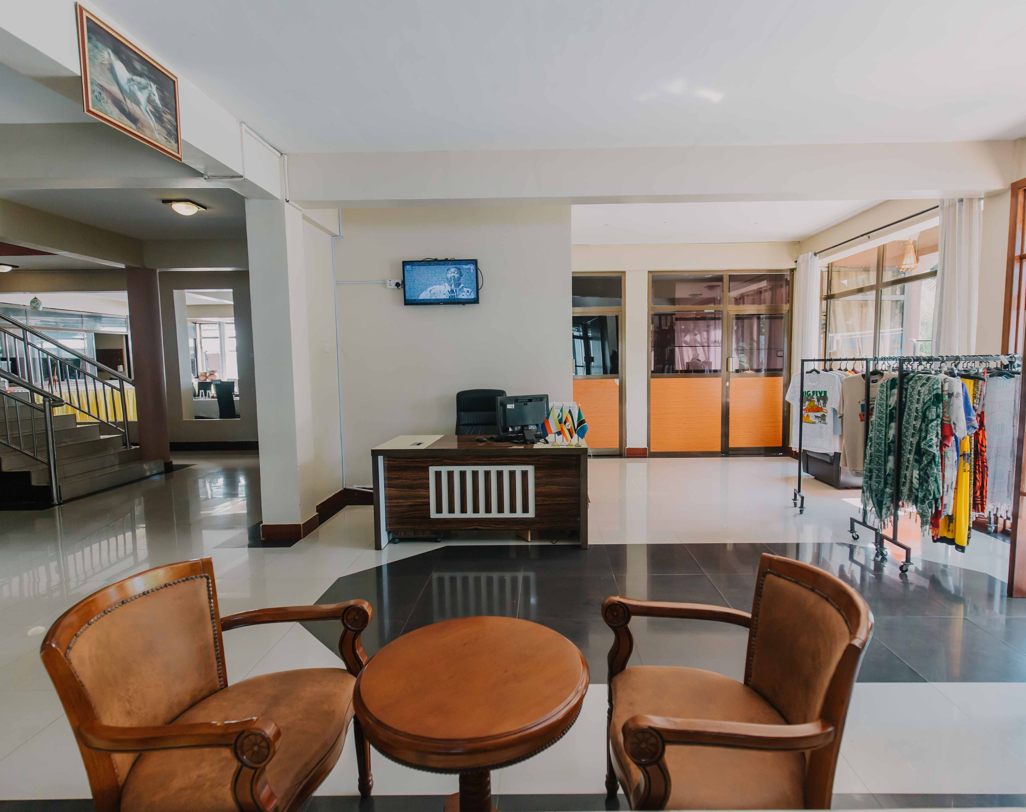 A hotel lobby with a reception desk, a television, a clothing rack, and two wooden chairs.