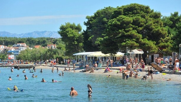 A beach with people swimming and sunbathing, a large tree, and buildings in the background. - Cosy apartment 5min to beach Mirela Borik (Zadar)