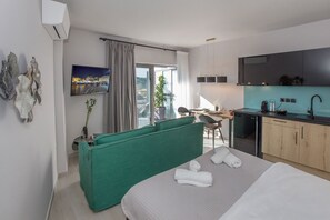 Ein modernes Hotelzimmer mit Bett, einem grünen Sofa, einem Schreibtisch mit Stuhl, einer Küchenzeile und einem Balkon mit Blick.