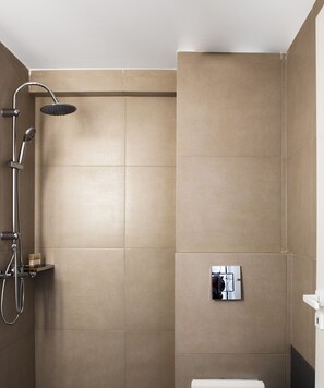 A modern bathroom with beige tiles, a shower with a handheld showerhead, a handheld showerhead, and a wall-mounted control panel. - Elysium Boutique Apartments (Zografos)